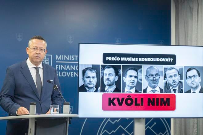 Finance Minister Ladislav Kamenický stands in front of a screen showing opposition politicians, whom he blames for the dire state of public finances and the need for consolidation.