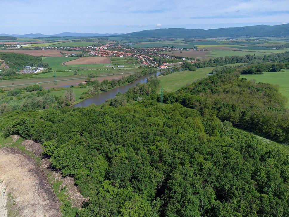 Tam kdesi pod stromami leží zakonzervovaná osada zo staršej doby bronzovej. Kameňolom si nenásytne ukrajuje z kopca a posúva sa čoraz bližšie. Za riekou je obec Kozárovce.