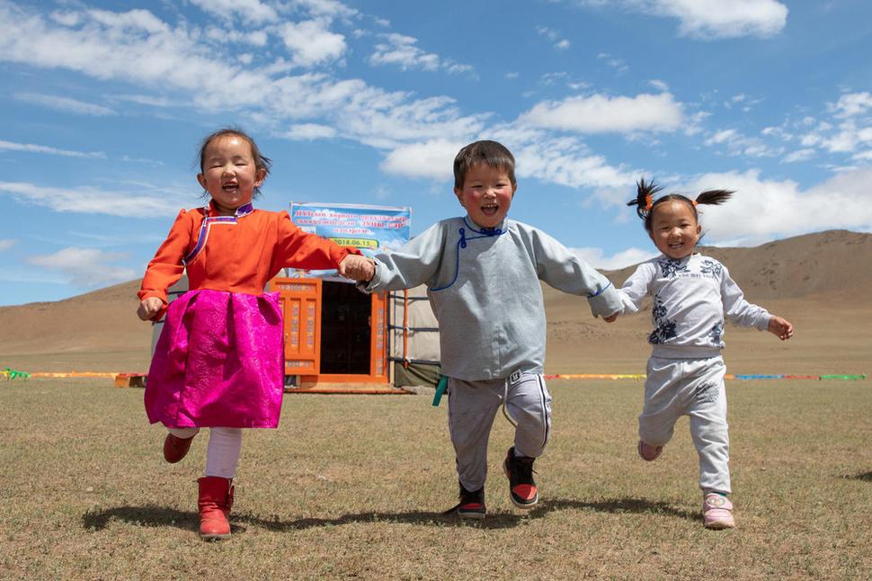 Tri smejúce sa deti v Mongolsku pred mobilnou škôlkou UNICEF, kde je deťom z nomádskych rodín počas roka poskytovaná predškolská starostlivosť a hravým spôsobom sa pripravujú na nástup do školy. Tiež majú možnosť stretávať sa tu s inými deťmi.