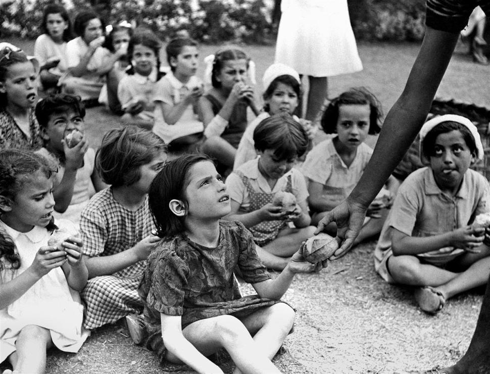 Deti usadené na zemi jedia čerstvé pečivo v jednom z letných táborov podporovaných UNRRA, predchodcom UNICEFu, Taliansko, cca 1945.