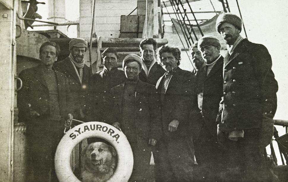 Sedem zachránených účastníkov podpornej skupiny so psom Oscarom na lodi Aurora. Zľava: Keith Jack, Alexander Stevens, Dick Richards, Ernest Wild, Irvine Gaze, Ernest Joyce, John Cope, Ernest Shackleton a kapitán John King Davis, január 1917.
