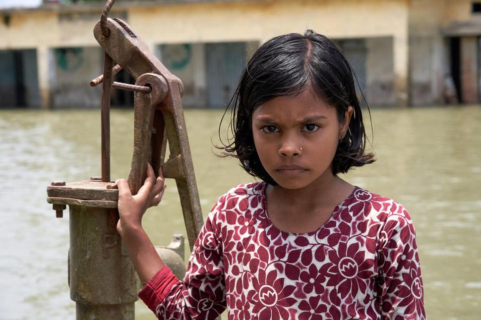Bangladéš, 2019: Ishrafi Khatun,10 stojí pred budovou školy, ktorá sa v dôsledku záplav ocitla pod vodou. Studňa, pri ktorej stojí, je jediným zdrojom pitnej vody v celej oblasti. V dôsledku silných dažďov bolo veľké územie Bangldéša zasiahnuté silnými záplavami.